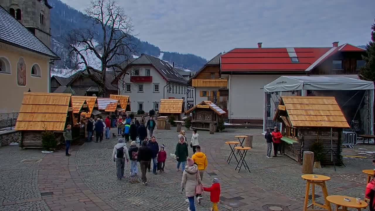 A cobblestone town square, under an overcast sky, features numerous small wooden market stalls surrounded by people, with a church and other buildings visible in the background.