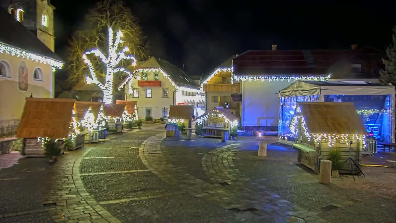A nighttime town square with small wooden structures decorated with lights, a large tree also lit up, and a stage under a tent.
