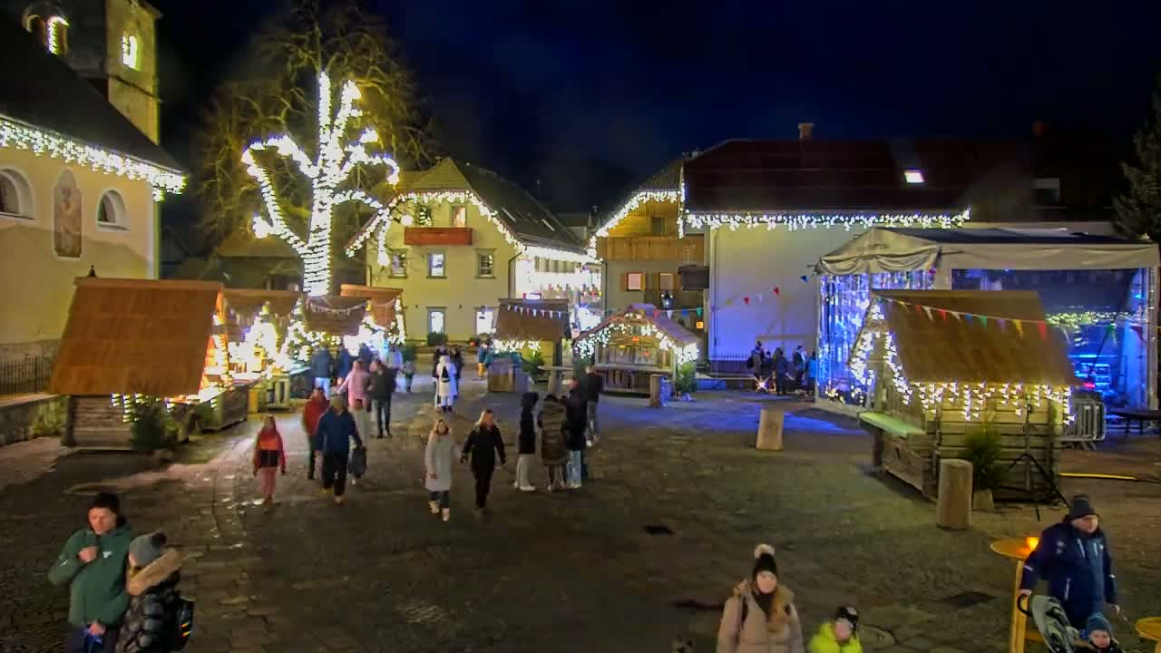 A nighttime outdoor Christmas market scene with wooden stalls decorated with lights, people strolling through the cobblestone square, and a large tree also illuminated with lights.