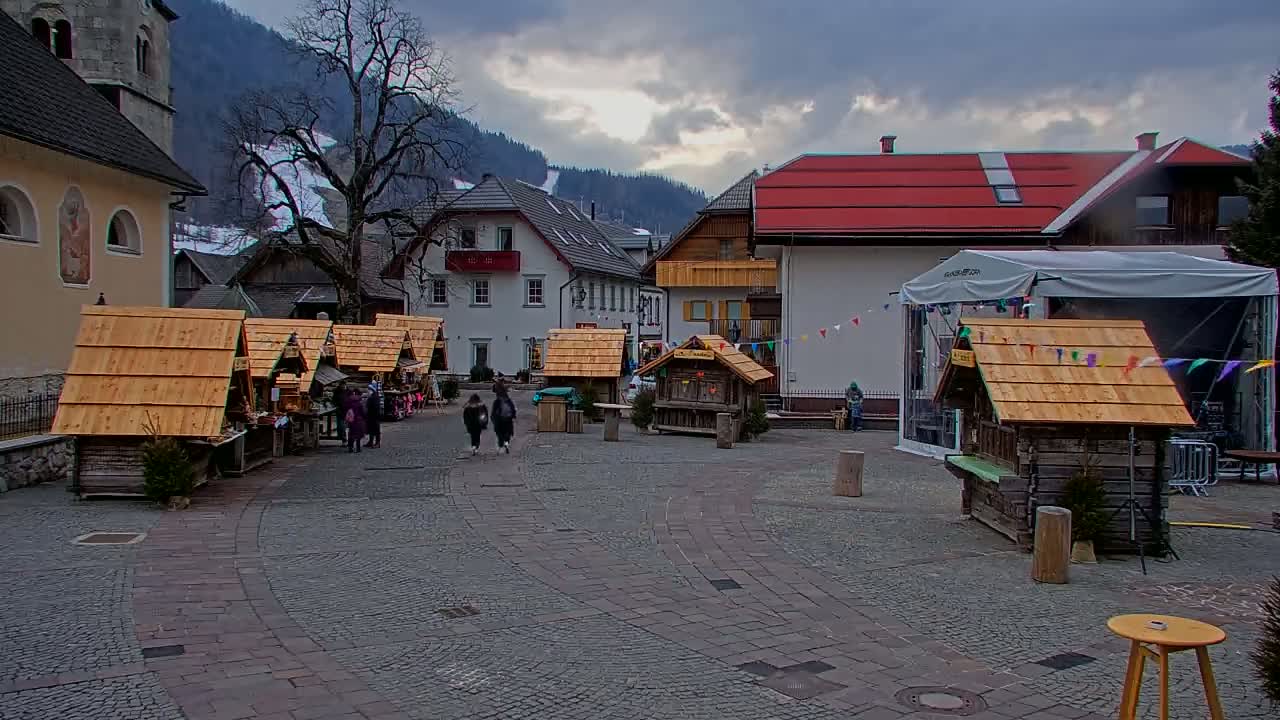 A cobblestone town square with several small wooden market stalls, surrounded by buildings and a mountain backdrop under a cloudy sky.