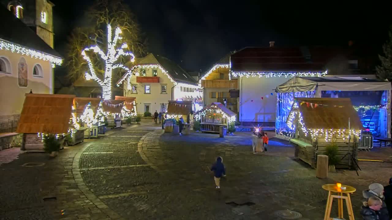A nighttime scene of a cobblestone town square with illuminated Christmas market stalls, a stage, and a large tree decorated with lights.