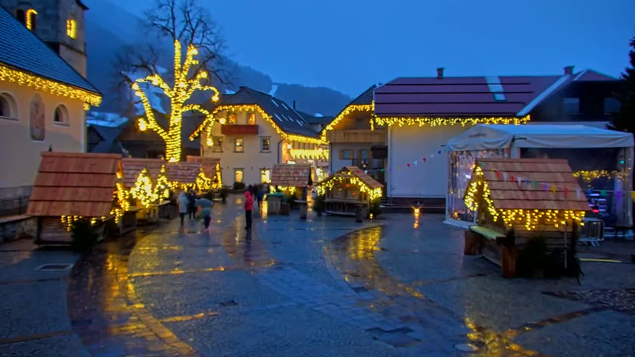 A wet cobblestone town square at night is illuminated by strings of warm-toned lights strung on buildings and small wooden market stalls, with a large tree also adorned with lights at its center.