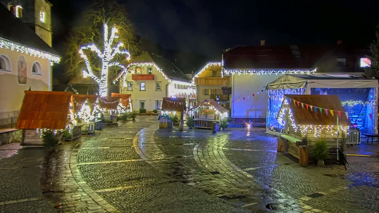 A cobblestone town square at night is decorated with Christmas lights strung on buildings and small wooden stalls, and it is wet from recent rain.