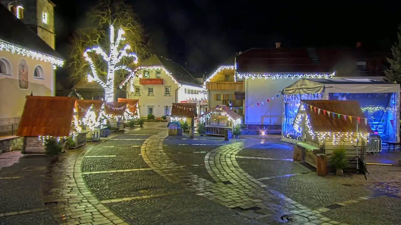 A cobblestone town square at night is decorated with numerous small wooden huts and string lights, illuminated against the backdrop of buildings adorned with festive lights.