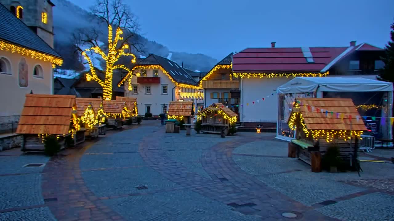 A town square at twilight is decorated with small wooden huts and string lights, and a large tree is also lit up with lights.