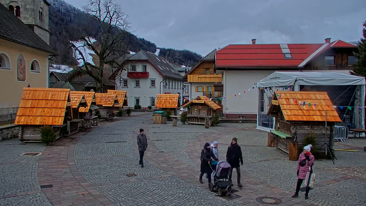 A town square with several small wooden stalls, a few people walking around, and buildings in the background under an overcast sky.