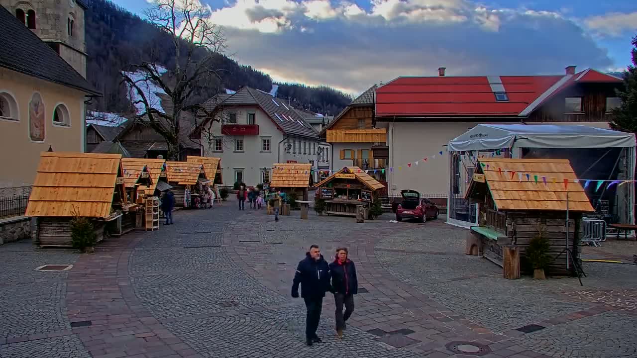 A cobblestone town square, under a partly cloudy sky, features several small wooden market stalls surrounded by buildings, including a church, with a couple walking in the center.