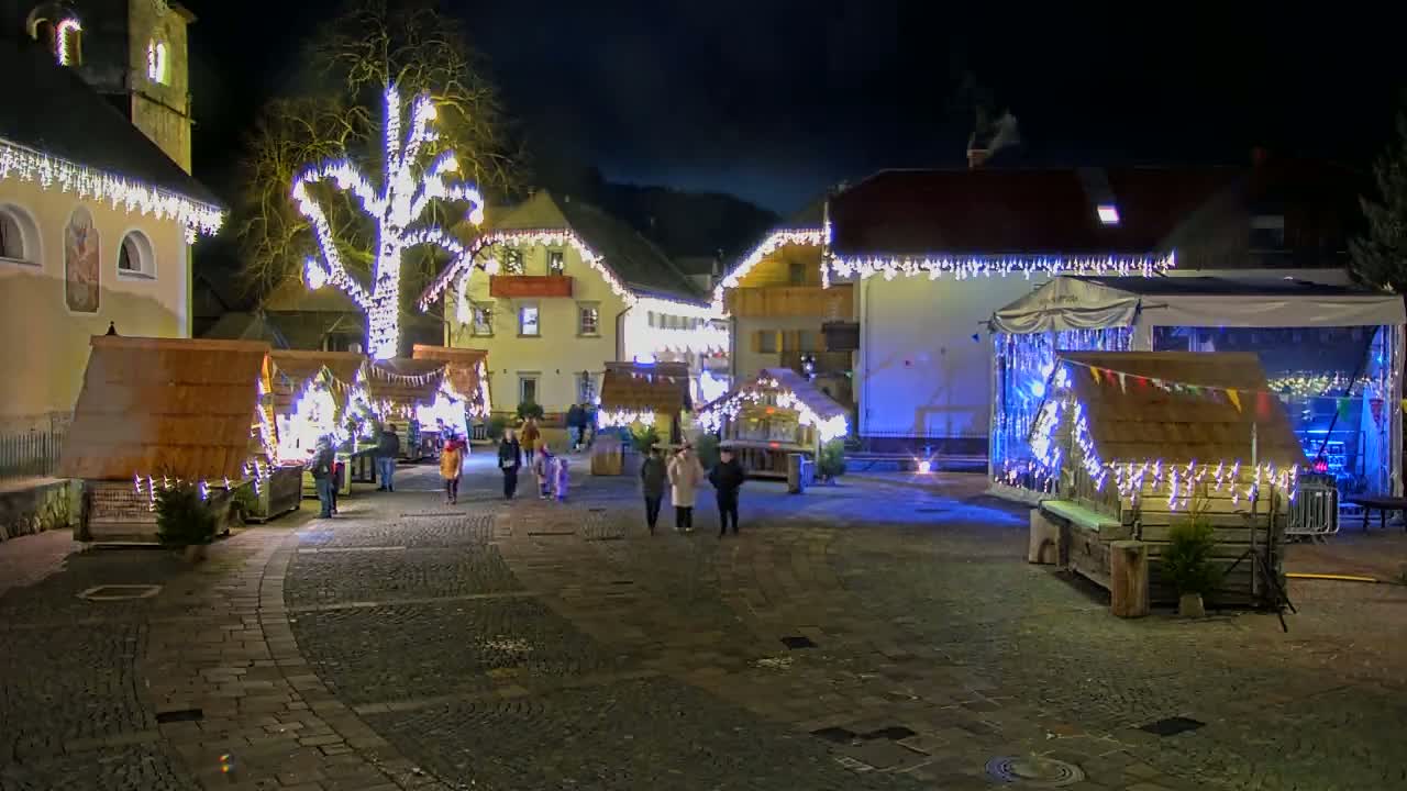 A nighttime outdoor Christmas market scene with small wooden stalls illuminated by string lights, a large tree also decorated with lights, and several people walking around.