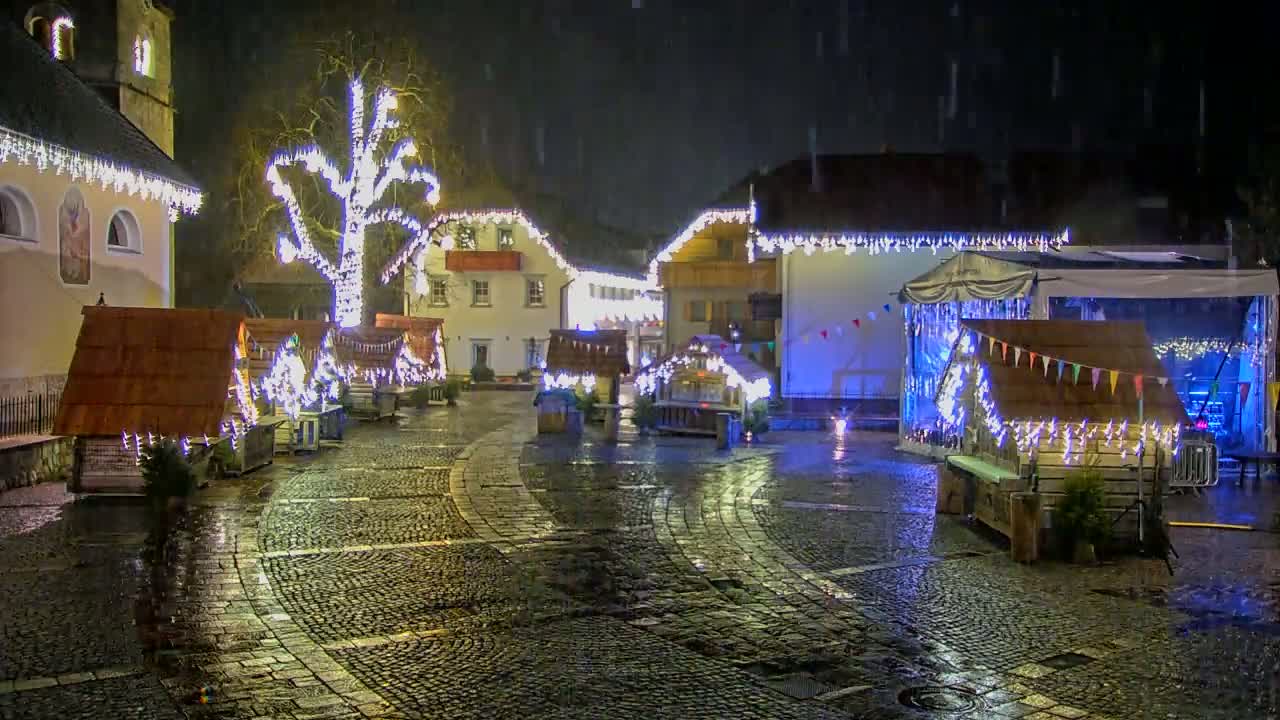 A cobblestone town square at night is decorated with Christmas lights and small wooden huts under a light rain.