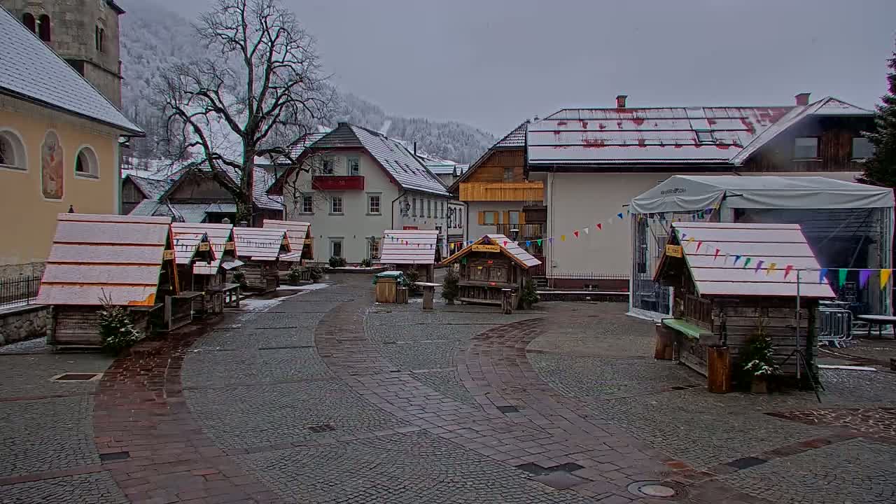 A snow-dusted village square features several small wooden stalls and buildings, with a larger building in the background under an overcast sky.