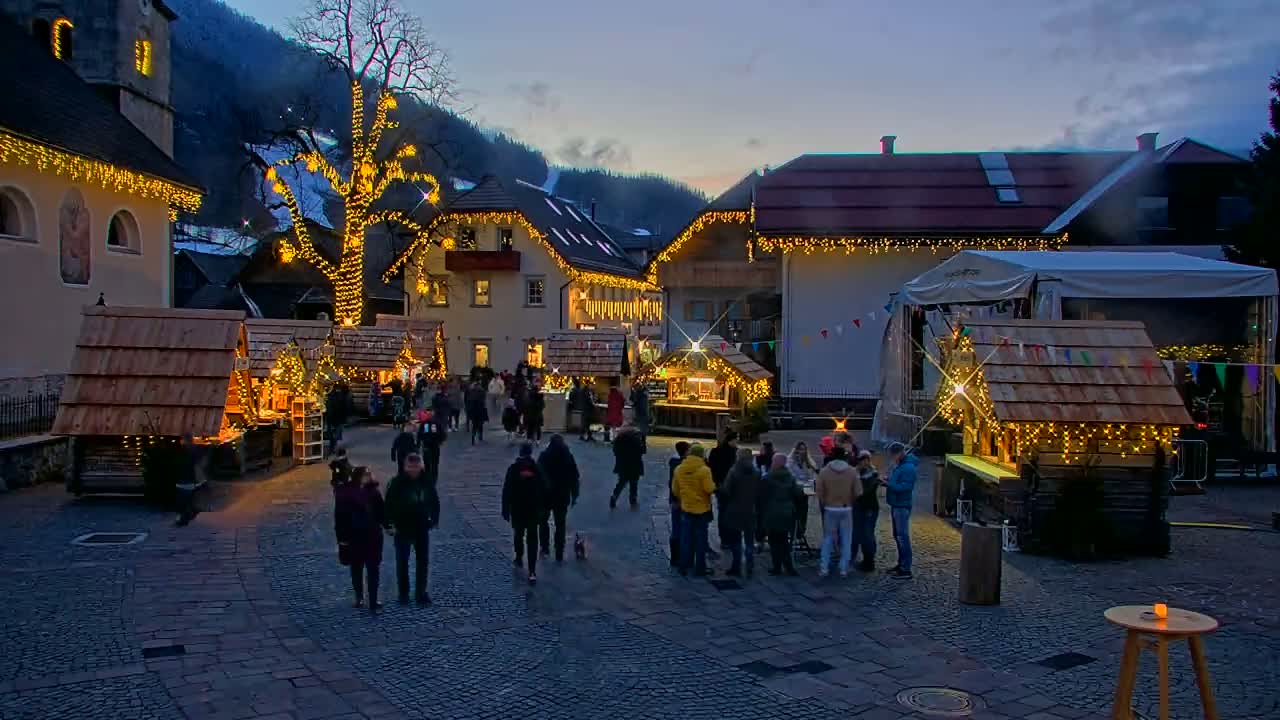 A nighttime outdoor Christmas market scene in a village nestled in a mountain valley, with wooden stalls illuminated by string lights and people milling about.