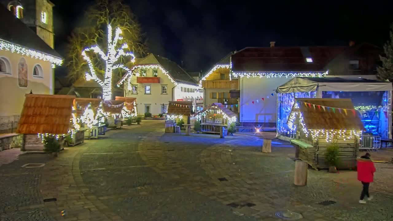 A town square at night is decorated with small wooden huts and strings of lights illuminating buildings and a large tree.