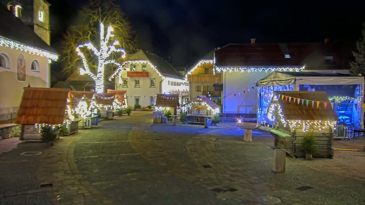 A nighttime scene of a town square decorated with numerous small wooden huts illuminated by strings of lights, a large tree also adorned with lights, and buildings with festive lighting.