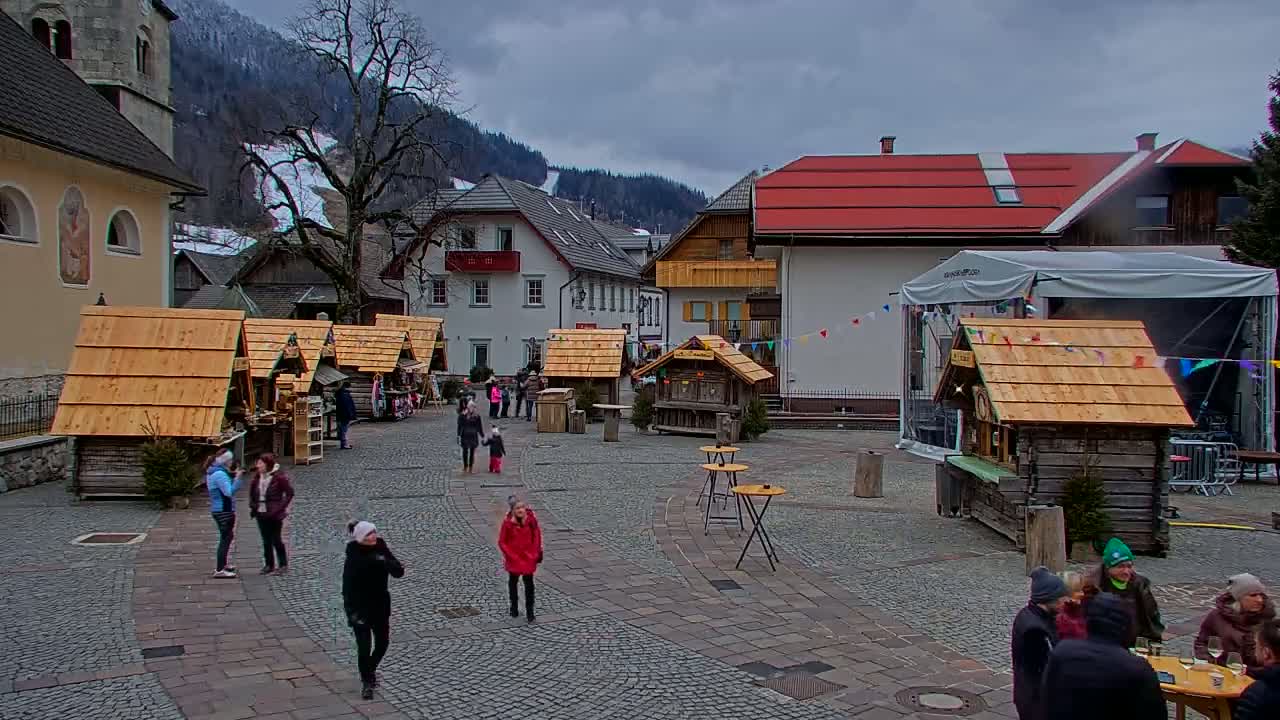 A cobblestone town square with several small wooden stalls, people milling about, and buildings lining the perimeter under an overcast sky.