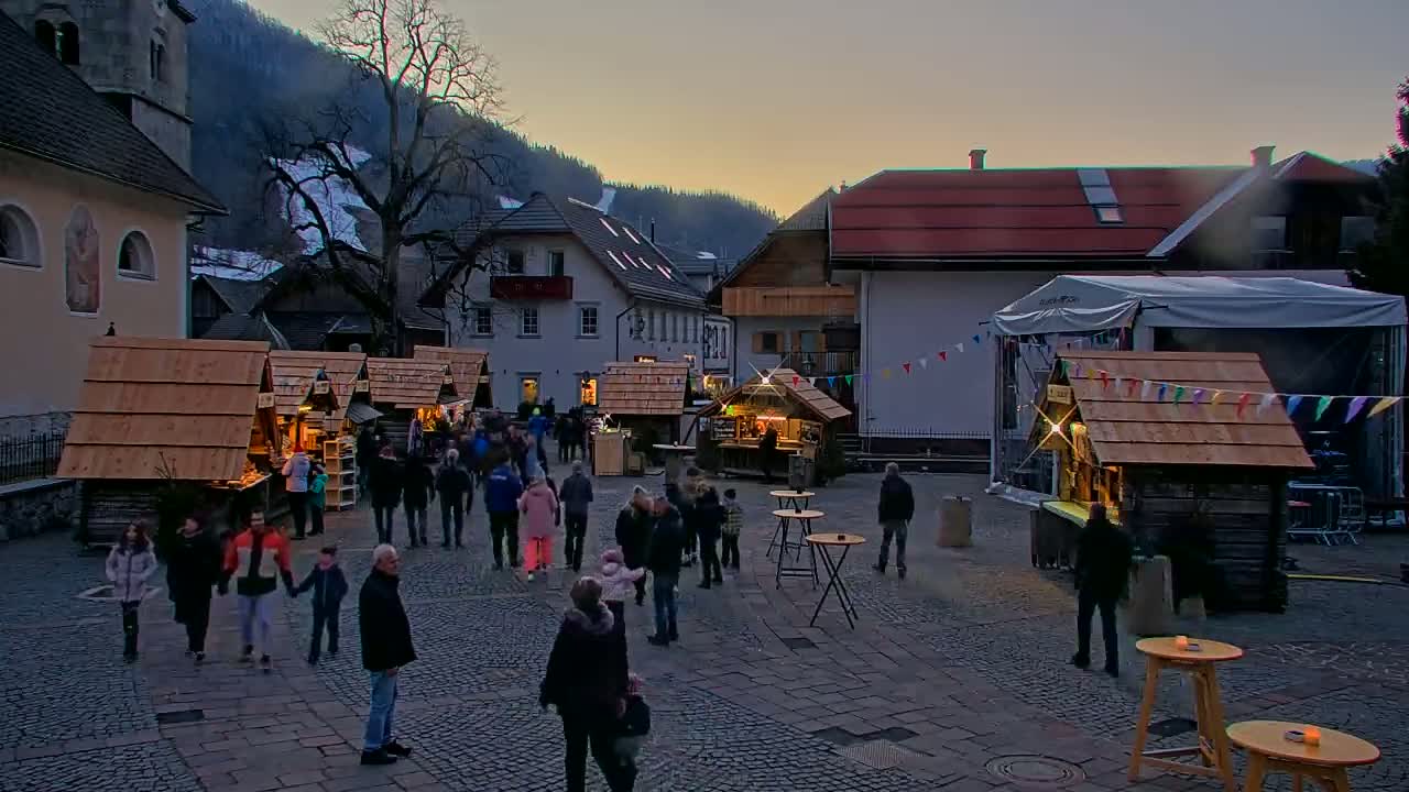 A town square at twilight, with several wooden market stalls surrounded by people, nestled between buildings in a mountain valley.