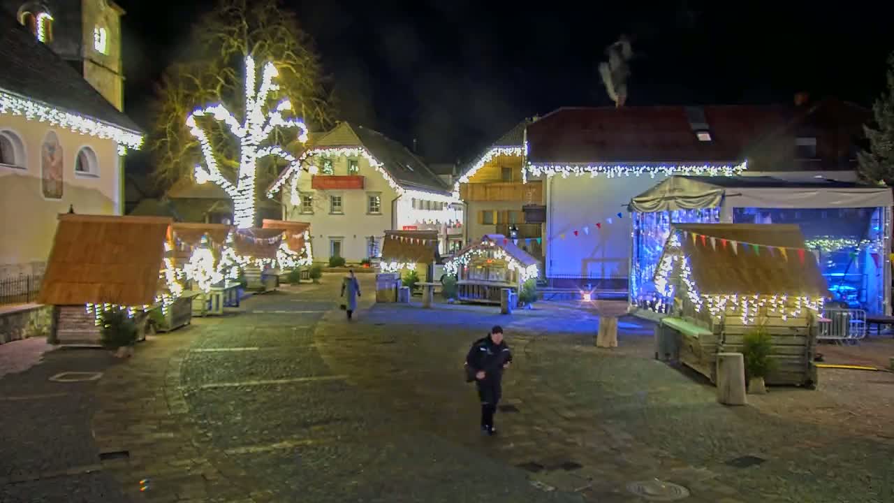 A nighttime scene of a town square decorated with Christmas lights and small wooden stalls, with a person walking in the cobblestone area.