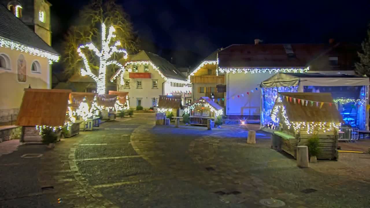 A town square at night, illuminated by strings of lights adorning buildings and small wooden market stalls.