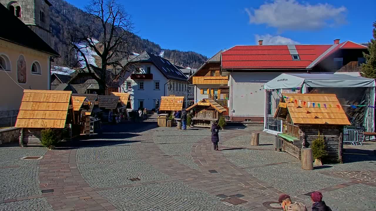 A cobblestone town square features several small wooden stalls, surrounded by buildings, under a mostly sunny sky.