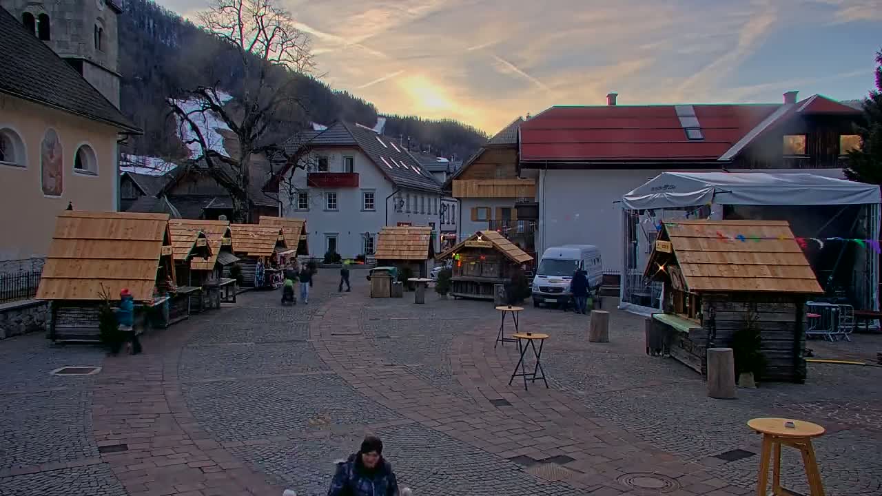 A cobblestone town square at sunset, featuring several wooden market stalls, buildings, a few people, and a van, is nestled against a snow-dusted mountainside.