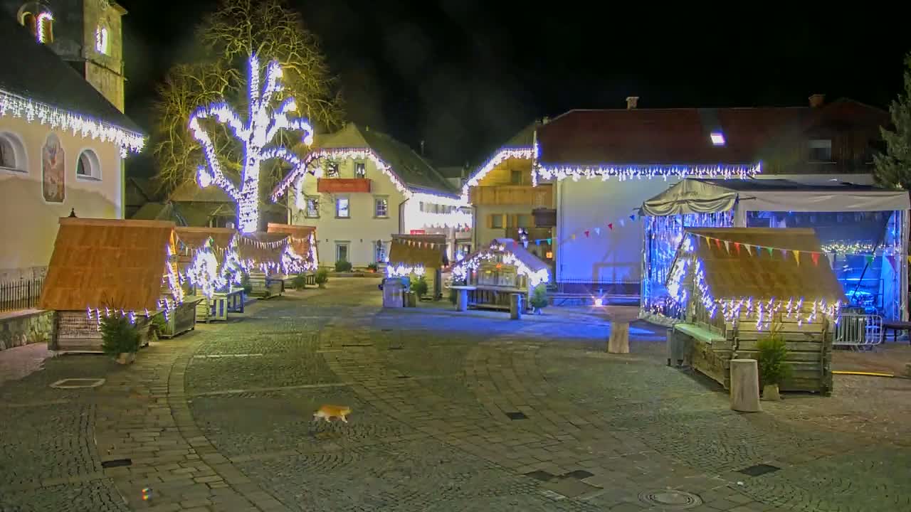 A town square at night is decorated with Christmas lights strung on buildings and small wooden stalls, and a cat walks across the cobblestone pavement.