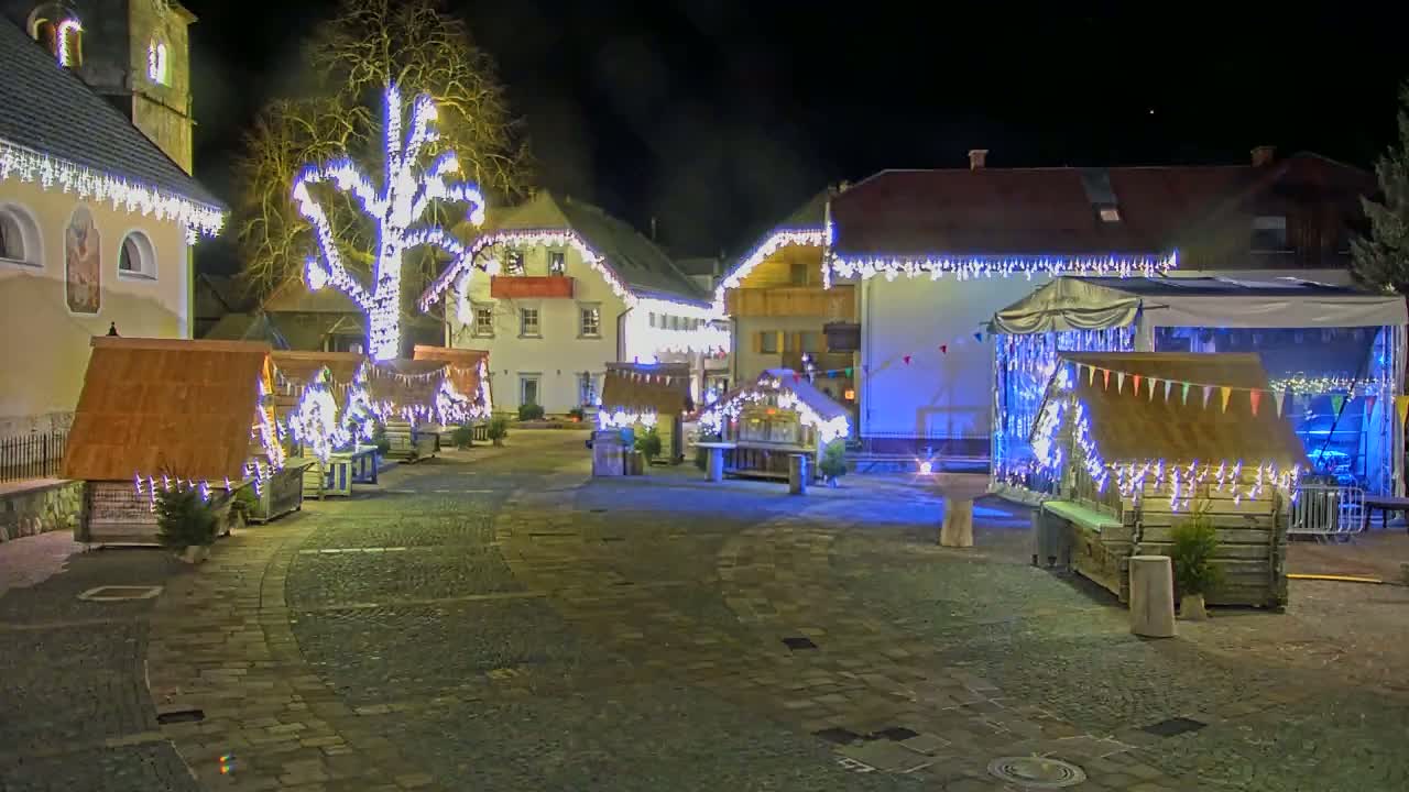 A nighttime outdoor scene of a town square decorated with Christmas lights and small wooden market stalls, centered around a large tree also adorned with lights.