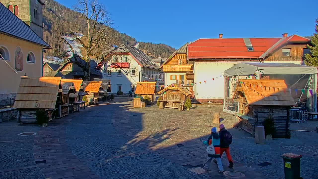 A sunny day in a European village square features several small wooden market stalls and buildings, with two people walking in the foreground.