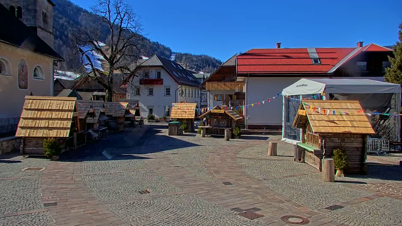 A cobblestone town square with several small wooden stalls, a few buildings, and a large tree, under sunny skies and mountains in the background.