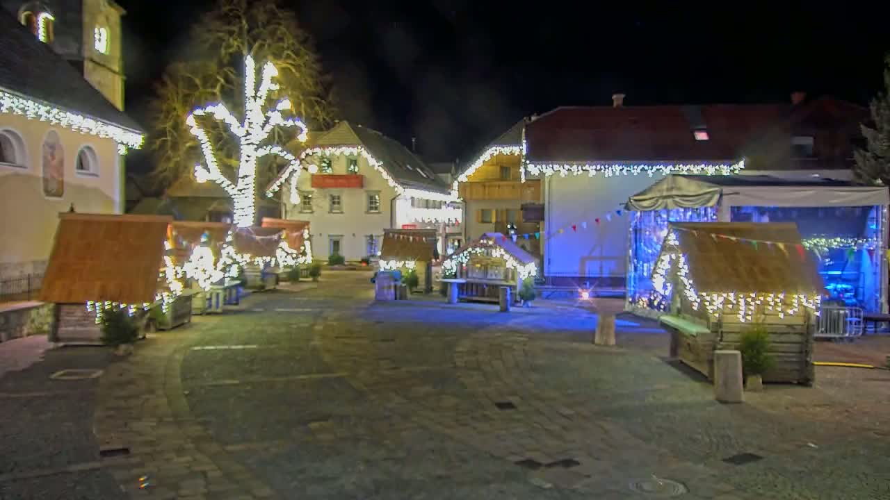 A nighttime scene of a town square adorned with twinkling lights on buildings and small wooden structures.