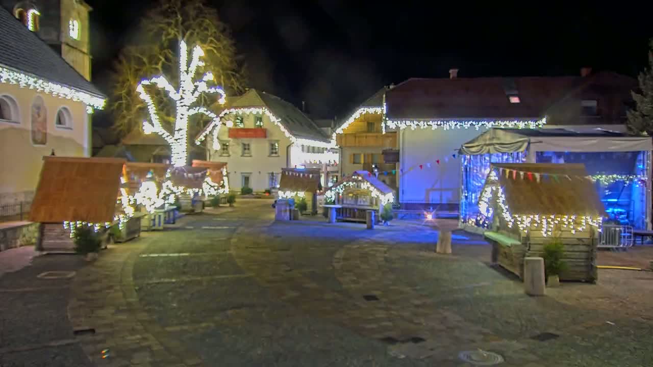 A nighttime scene of a town square with numerous small wooden huts decorated with strings of lights, situated around a large tree also adorned with lights.