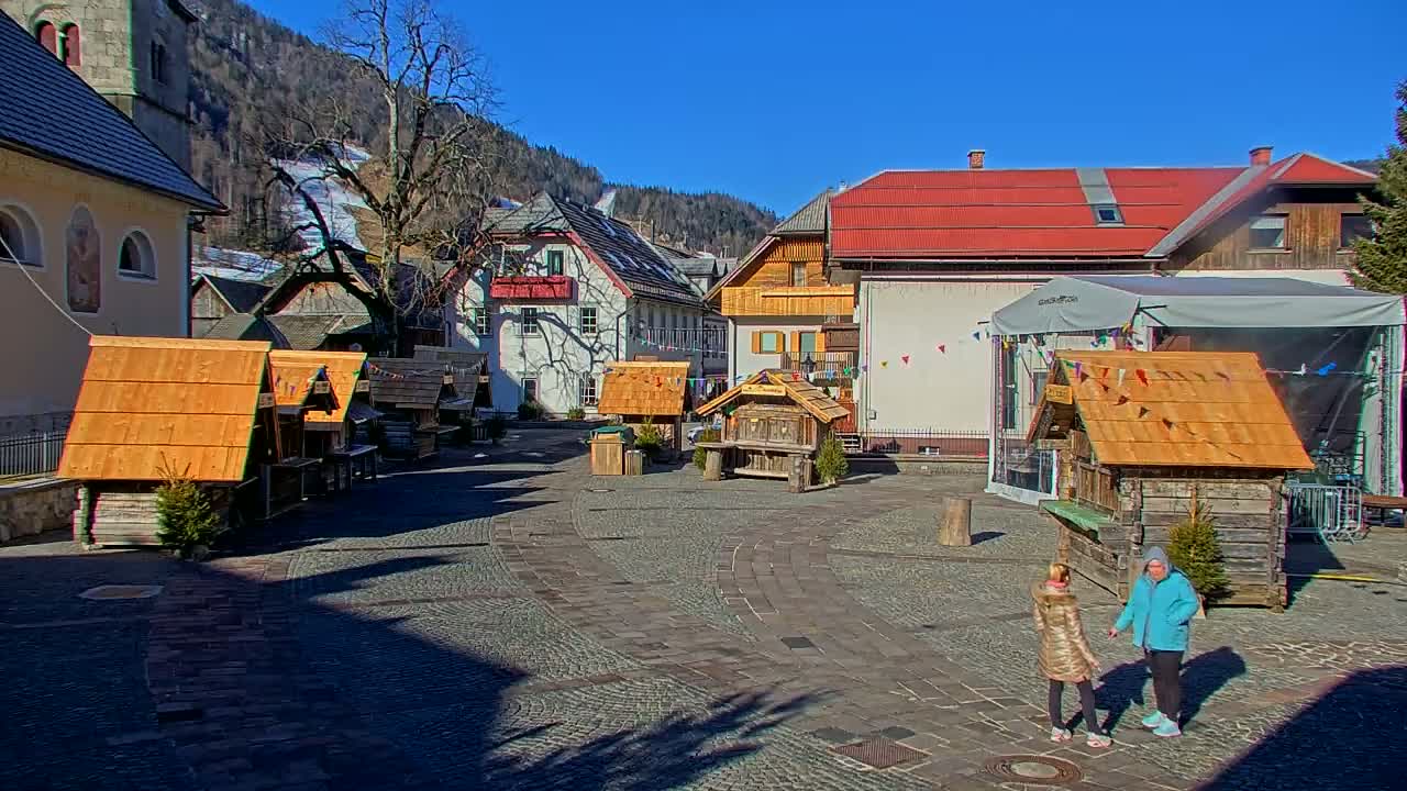 A cobblestone town square with several small wooden stalls, surrounded by buildings, under a clear blue sky on a sunny day with snow visible on a hillside in the background.