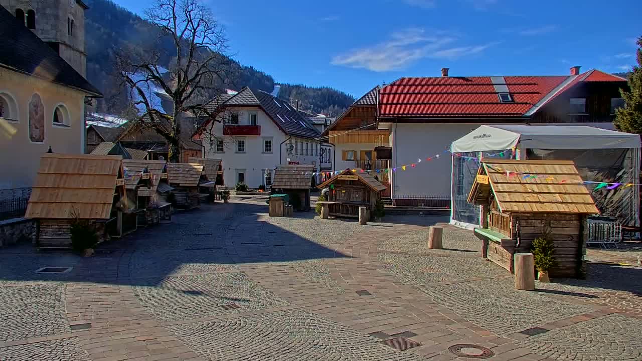 A cobblestone town square with several small wooden market stalls, surrounded by buildings and mountains under a clear, sunny sky.