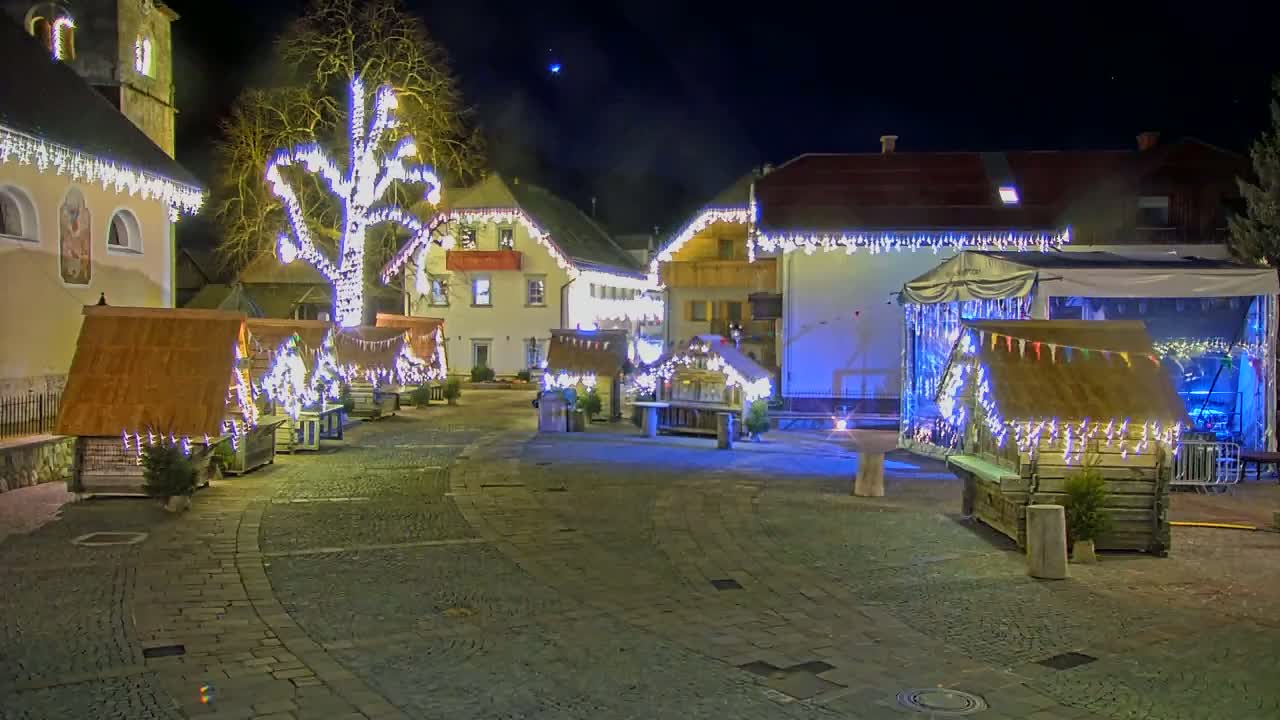 A nighttime scene of a village square with several small wooden huts decorated with lights, a large tree also lit up, and buildings adorned with holiday lights.