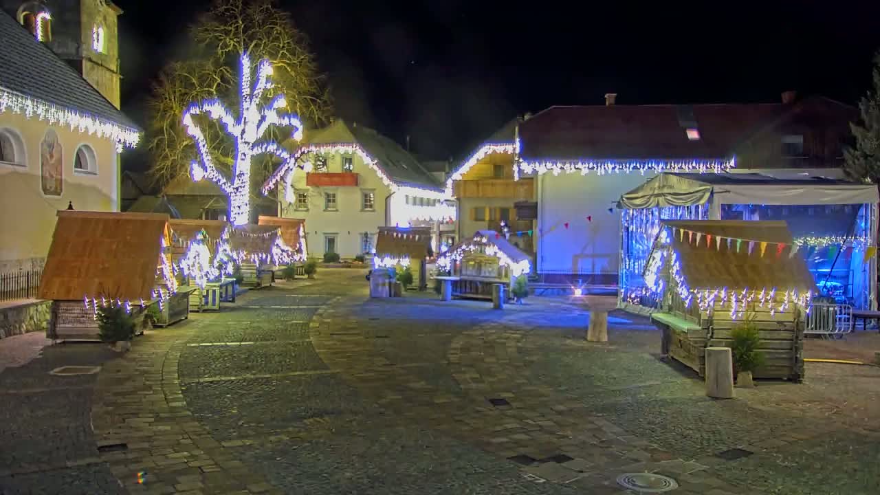 A nighttime scene of a town square decorated with Christmas lights and small wooden stalls, surrounding a large tree also lit up.