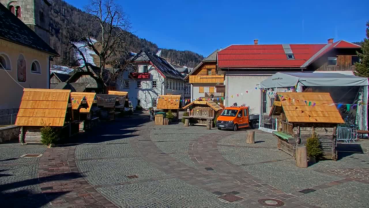 A sunny day in a village square with several small wooden stalls, a few buildings, and an orange truck, all situated against a backdrop of snow-dusted mountains.