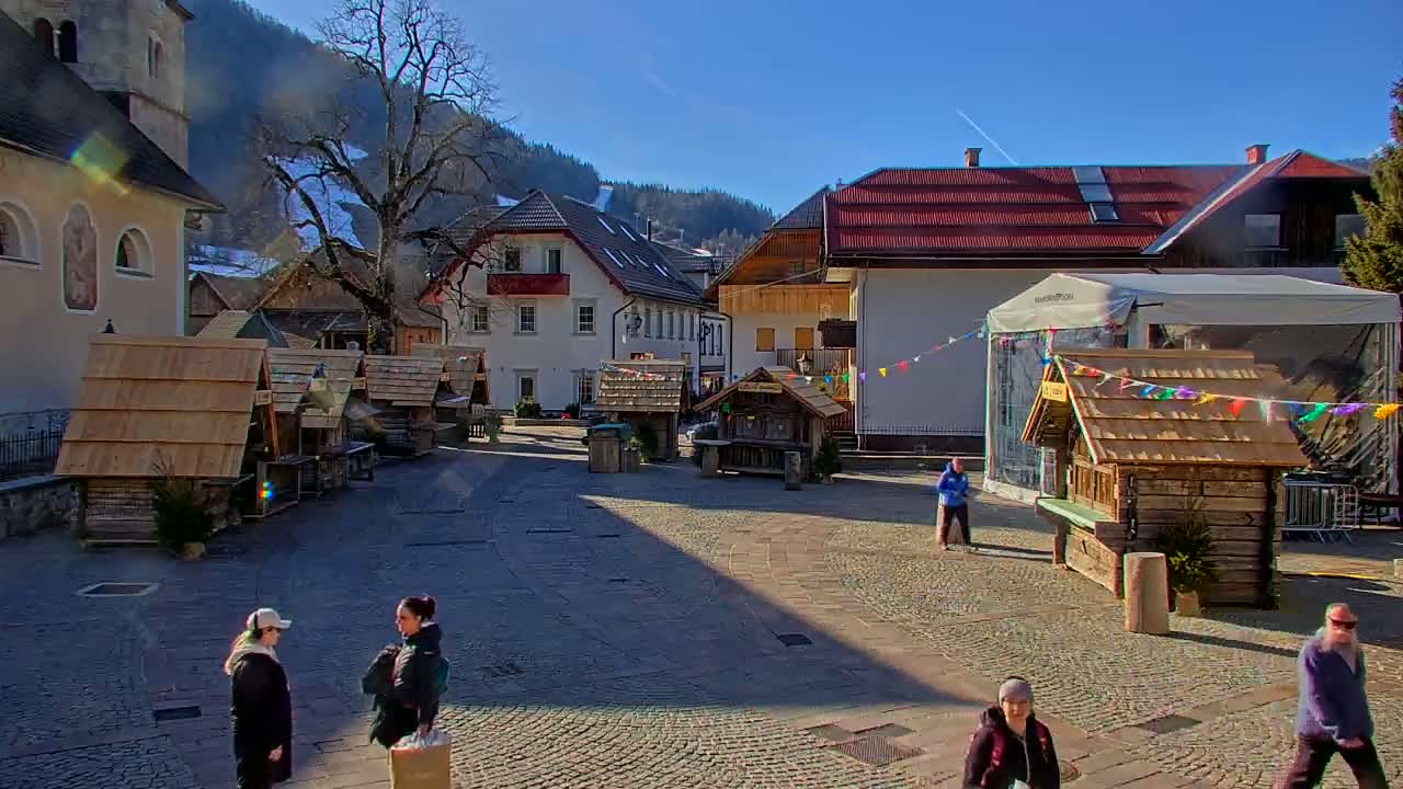 A sunny day in a European village square with several small wooden market stalls, buildings with red roofs, and a few people walking around.