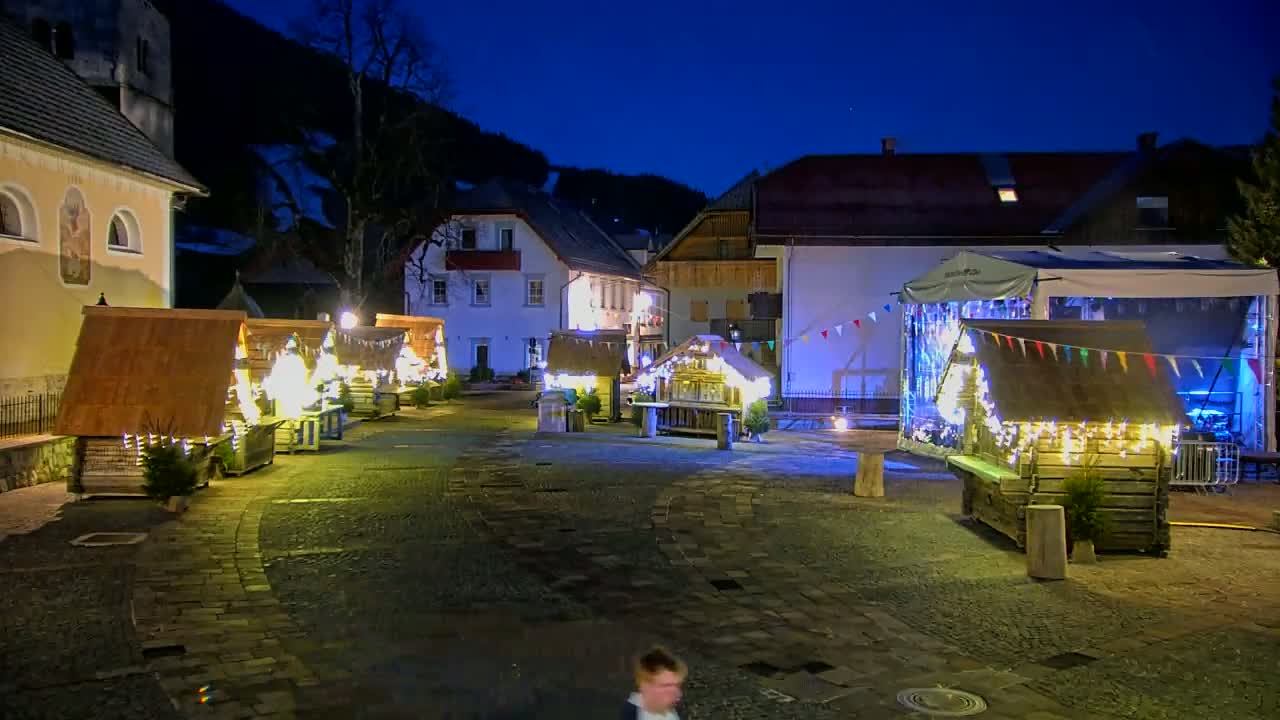 A cobblestone town square at night is illuminated by string lights on several small wooden market stalls and buildings, with a dark mountain visible in the background under a twilight sky.