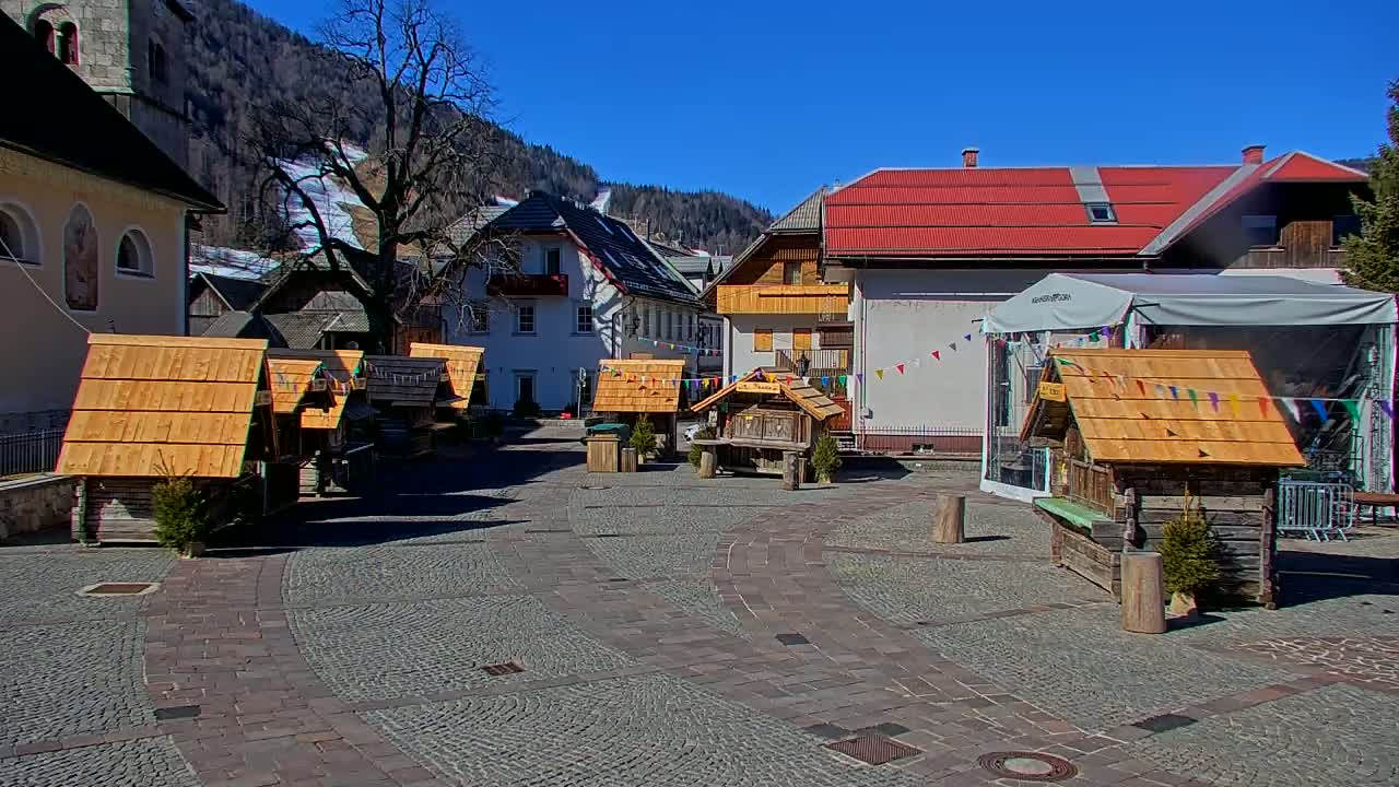 A cobblestone town square in a mountainous region features several small wooden stalls under a sunny sky.