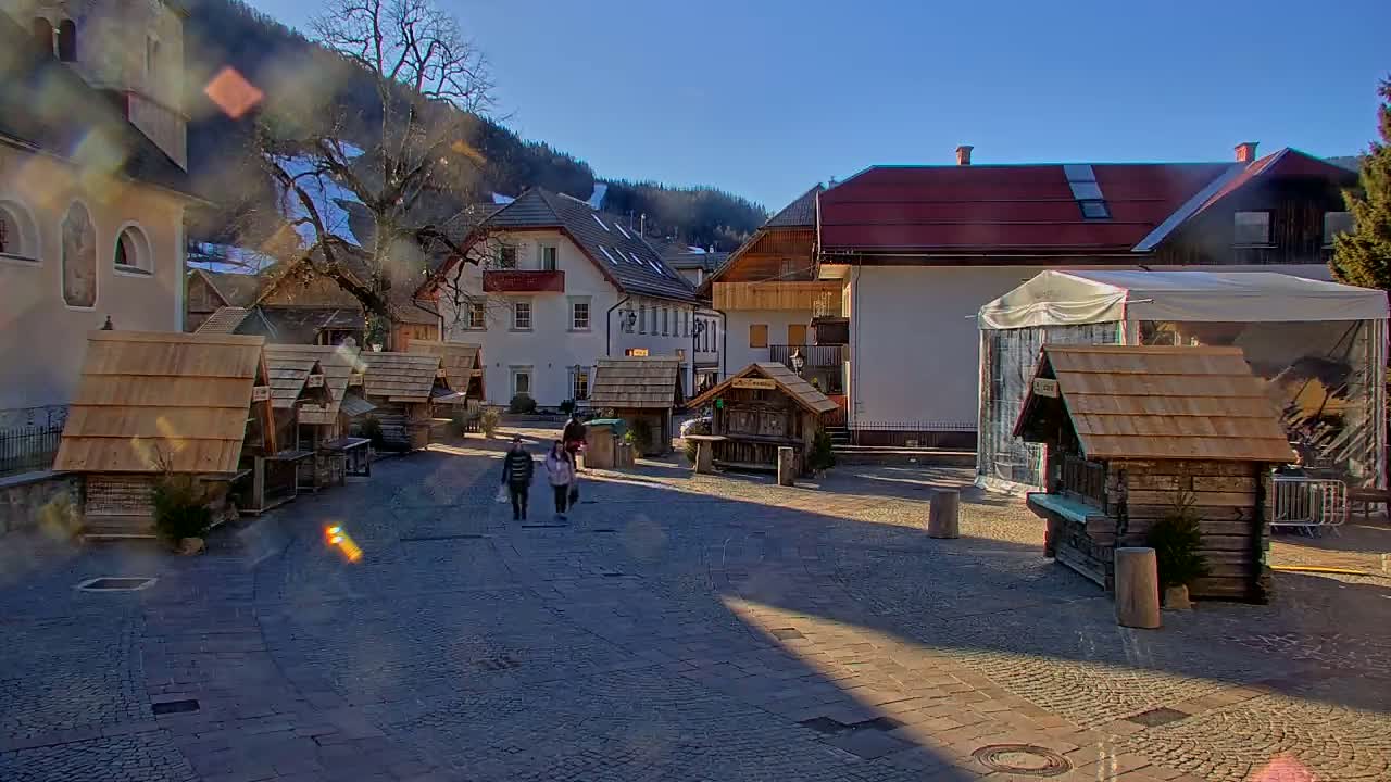 A sunny day in a European village square shows several small wooden market stalls and buildings, with a few people walking.