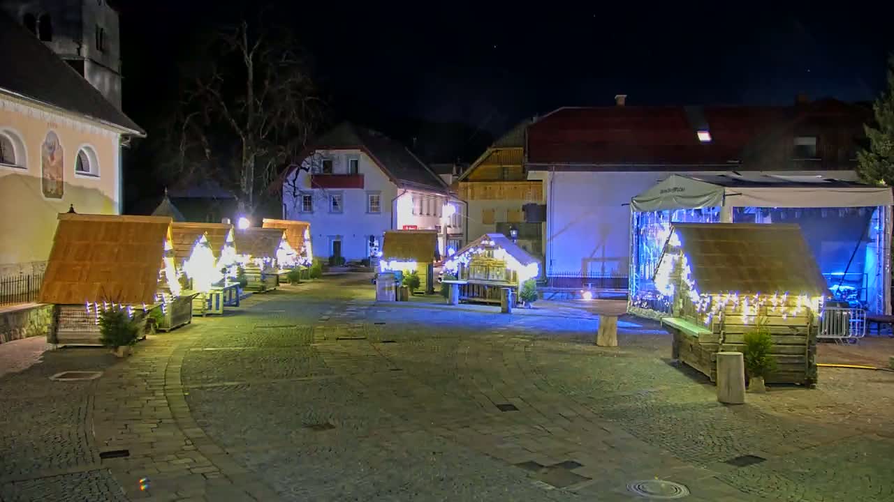A nighttime view of a town square with several small, illuminated wooden structures, possibly market stalls or holiday displays, surrounded by buildings under a clear, dark sky.