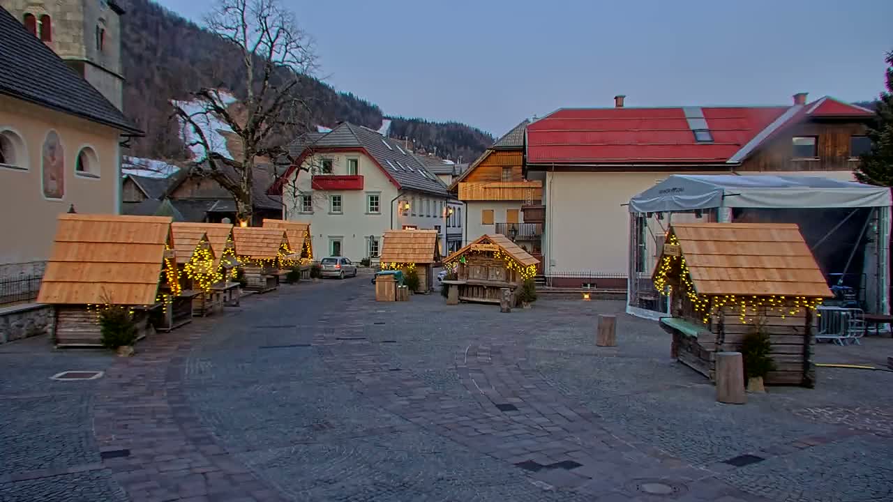 A cobblestone town square at dusk, features several small wooden market stalls decorated with lights, nestled among buildings against a backdrop of snow-dusted mountains.