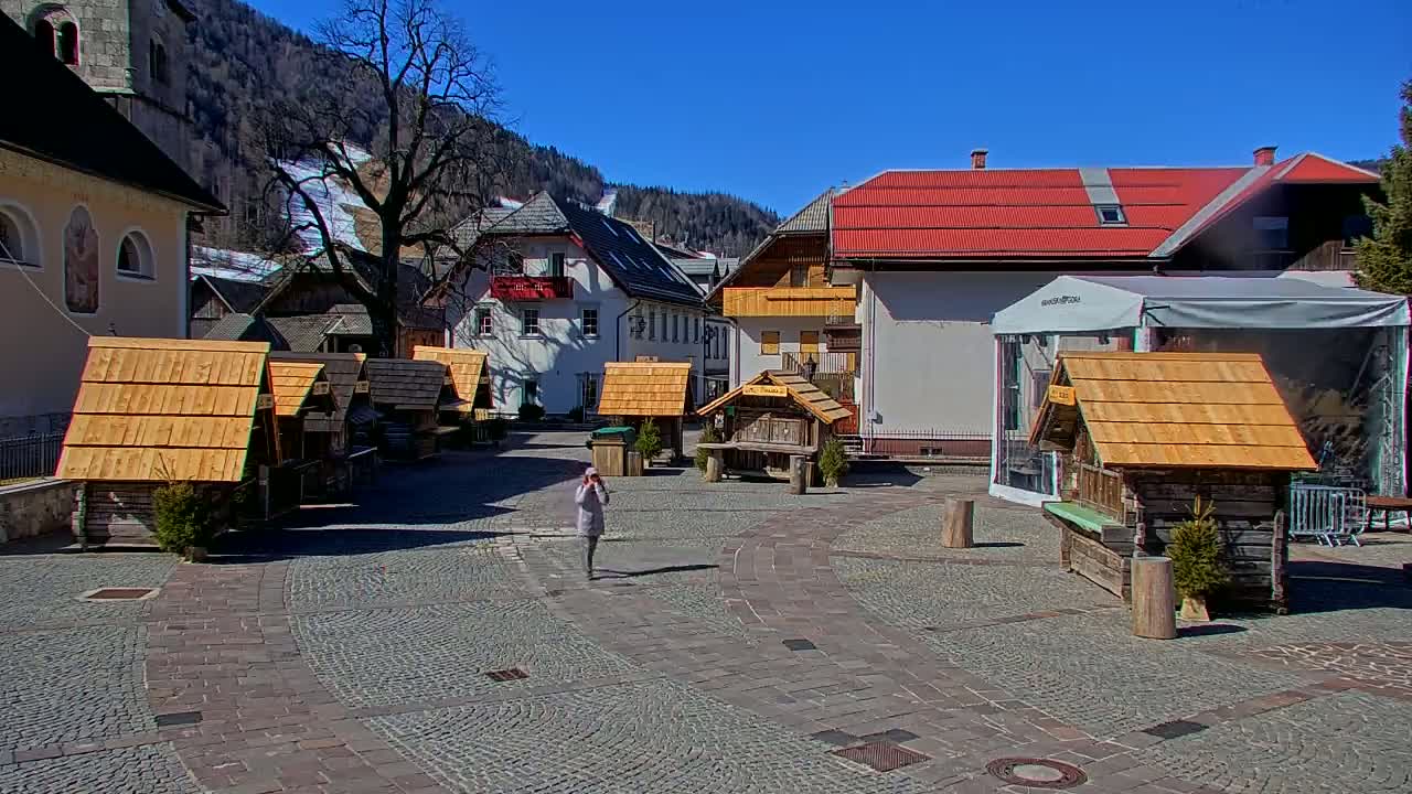 A person walks through a cobblestone town square with several small wooden structures and buildings lining the perimeter, under a sunny sky.