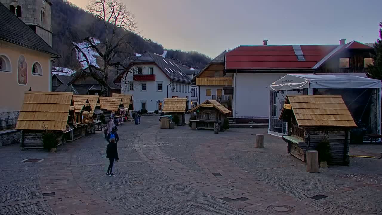 A cobblestone town square with several small wooden market stalls, a few people, and buildings including a church, under a dusky sky with mountains in the background.