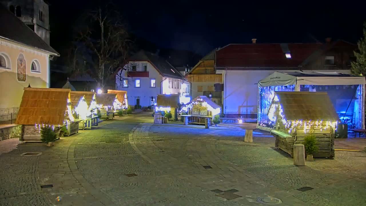 A nighttime scene of a town square with several small wooden huts decorated with lights, situated near buildings under a clear, dark sky.