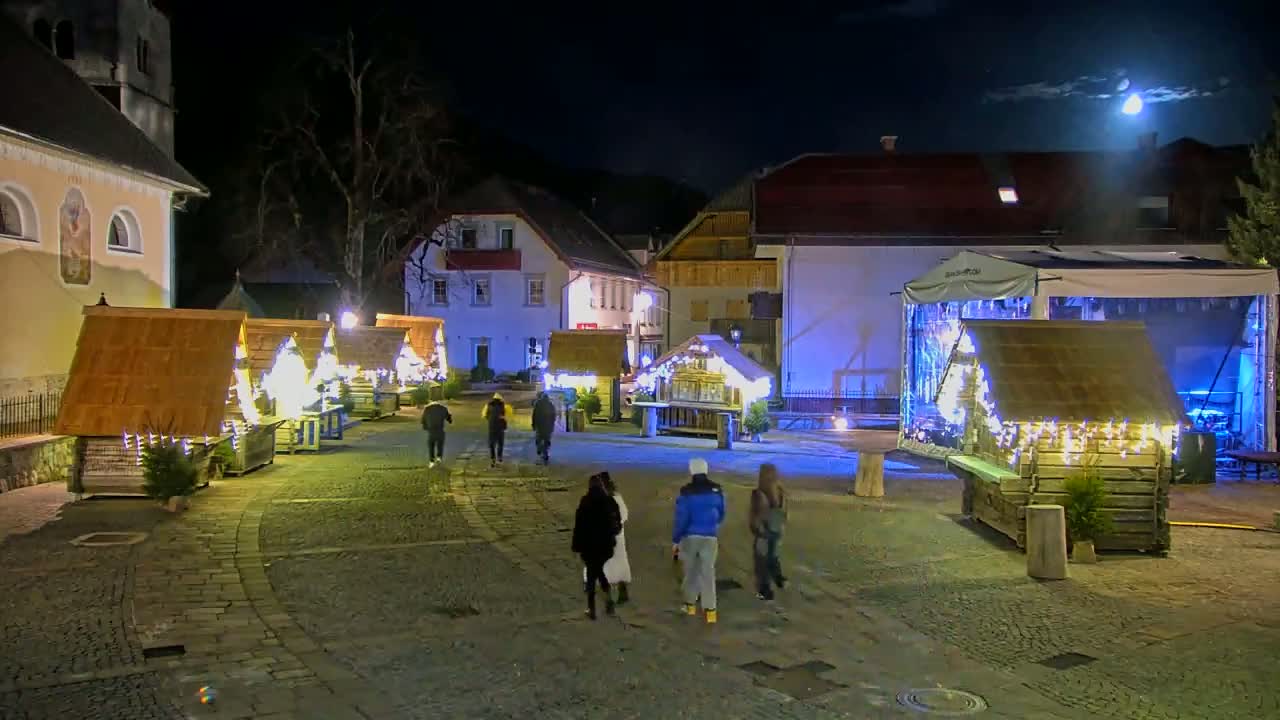 A nighttime outdoor scene shows a town square with several illuminated wooden market stalls, a stage, and a few people walking around under a mostly clear sky with a bright moon.