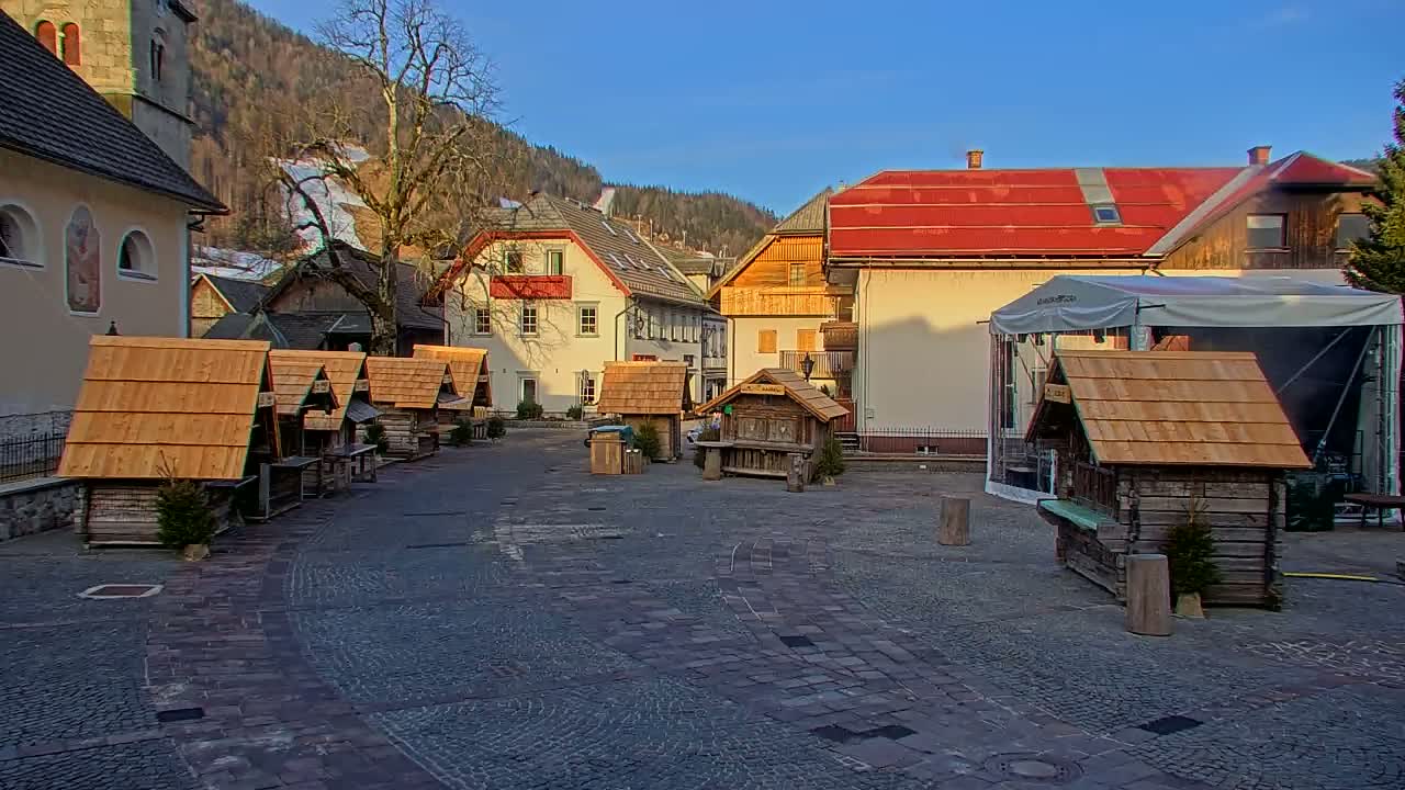 A cobblestone town square, surrounded by buildings and small wooden stalls, under a sunny sky with mountains in the background.
