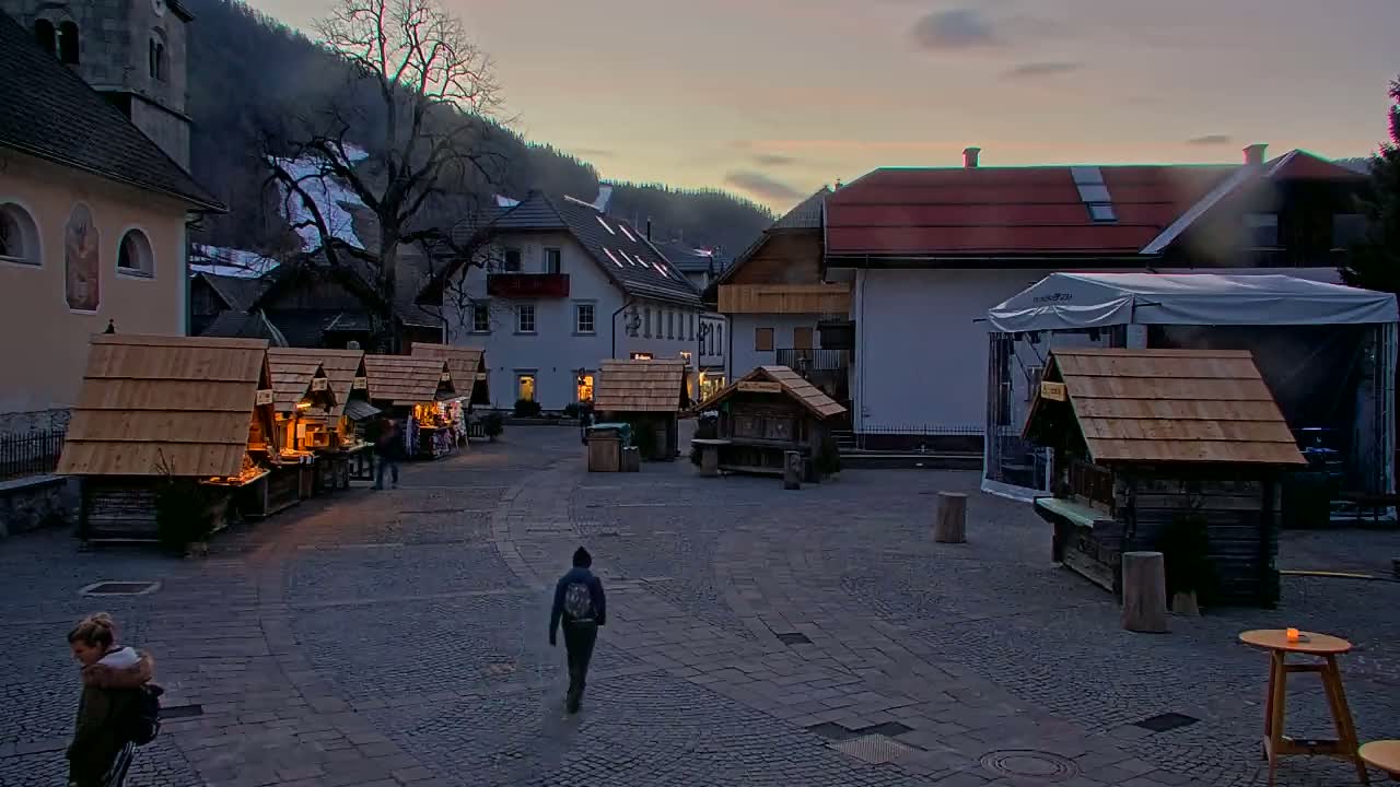 A cobblestone town square at dusk, nestled against a mountainside, features several small wooden market stalls and a few people walking around.