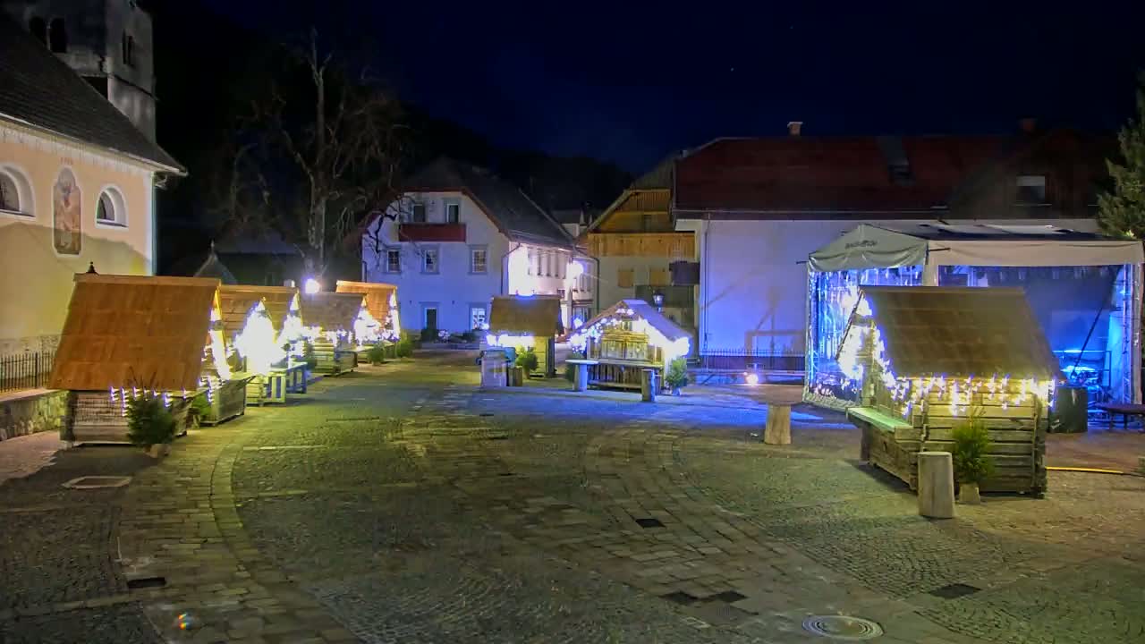 A nighttime scene of a cobblestone town square with several illuminated wooden market stalls and buildings.