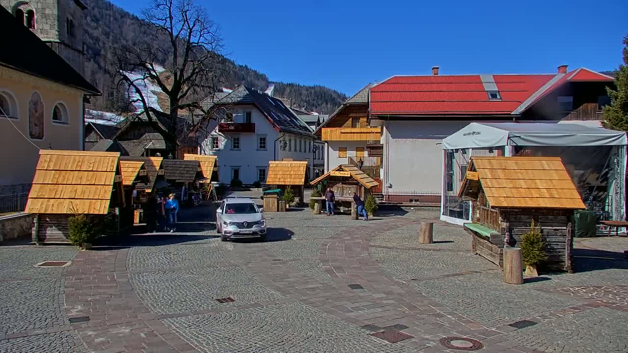 A village square with several small wooden stalls, a car, and a few people, under a clear blue sky in sunny weather, is situated against a backdrop of snow-covered mountains.