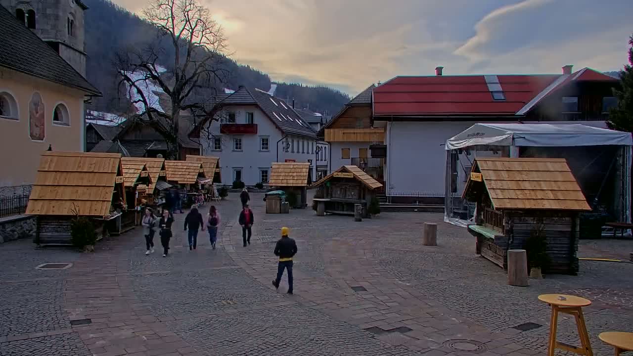 A town square with several small wooden stalls, a few people walking, and buildings in the background under a partly cloudy sky.