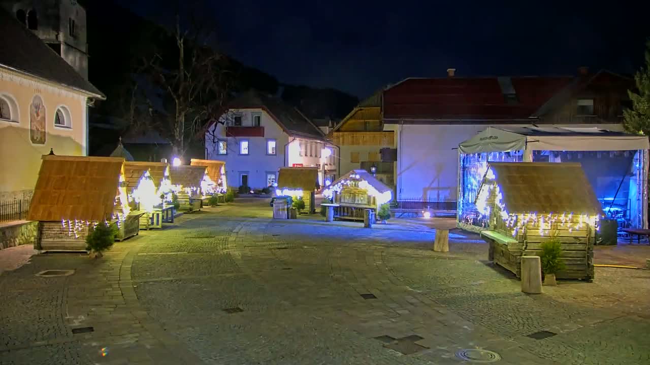 A nighttime outdoor scene shows several small wooden structures decorated with string lights in a cobblestone town square, with buildings in the background.